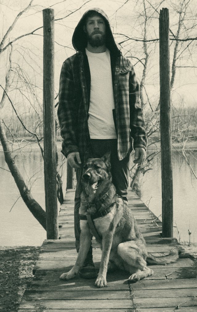 Ty McDuffey standing on a wooden footbridge over a river with his German shepherd, rendered in ivory on bottle green